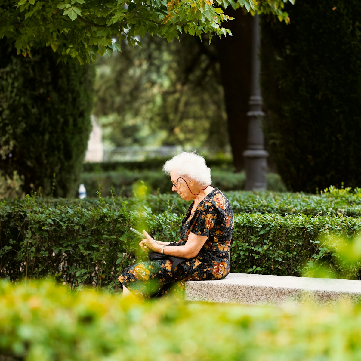 An elderly woman using a smartphone while sitting on a stone bench in a garden